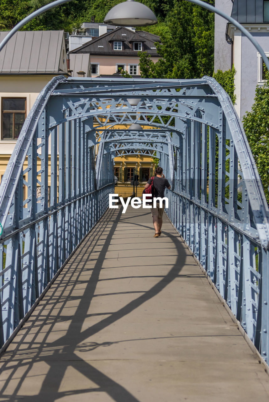 REAR VIEW OF MAN WALKING ON FOOTBRIDGE AGAINST BUILDING