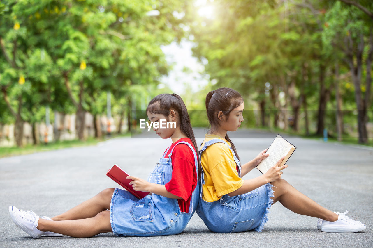 Side view of sisters reading book while sitting on road