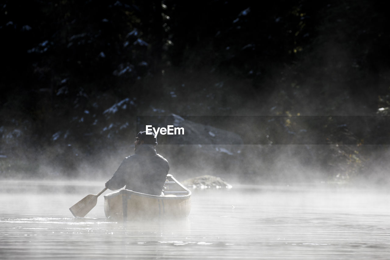 Man with hat holds paddle and canoes in fog on secluded lake and takes in the view of nature