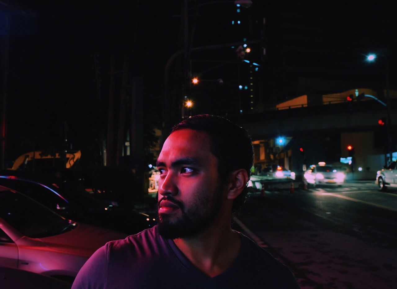 Close-up of man looking away while standing on street in illuminated city at night