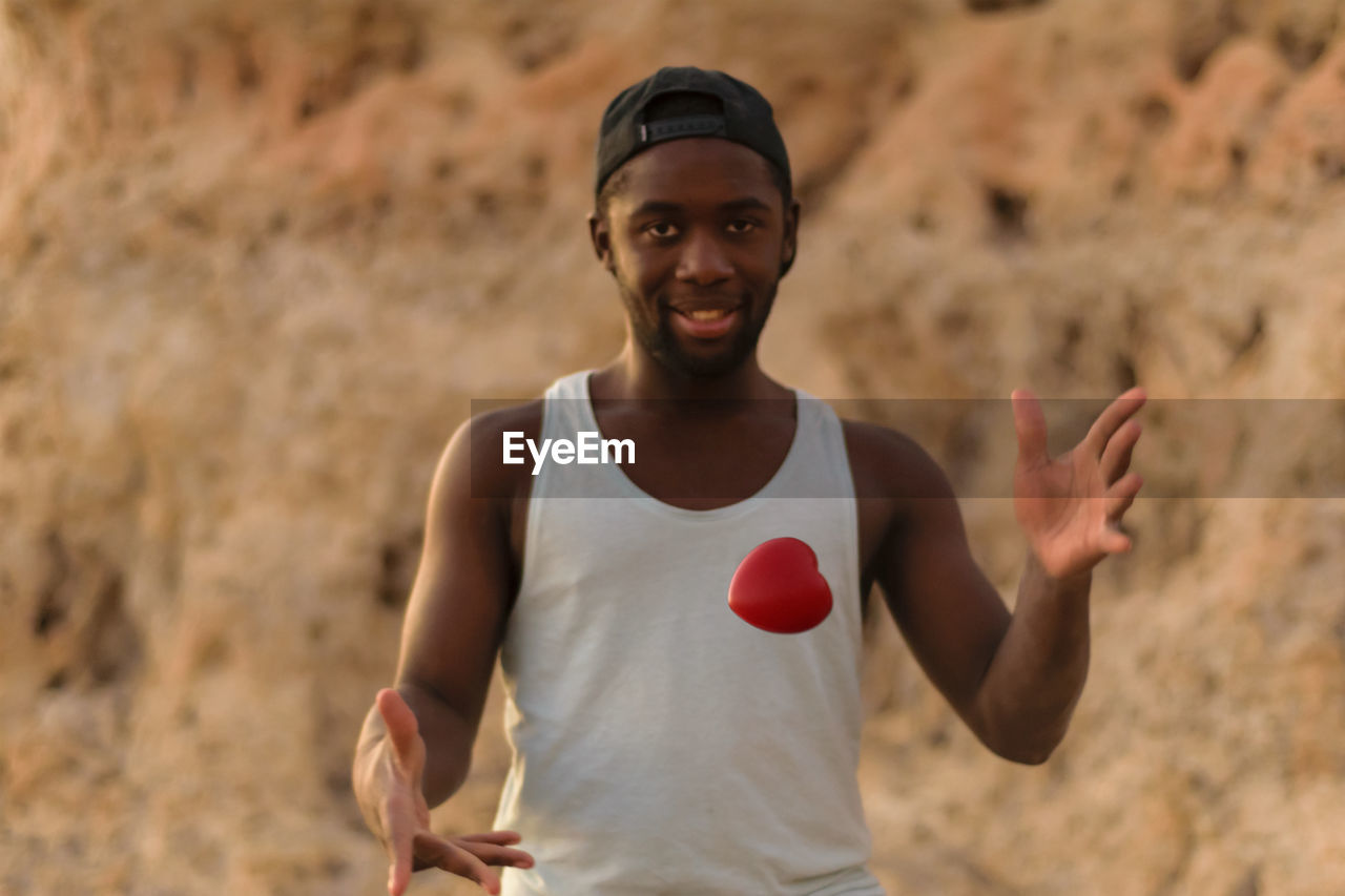 Portrait of young man catching red heart shape against rock formation
