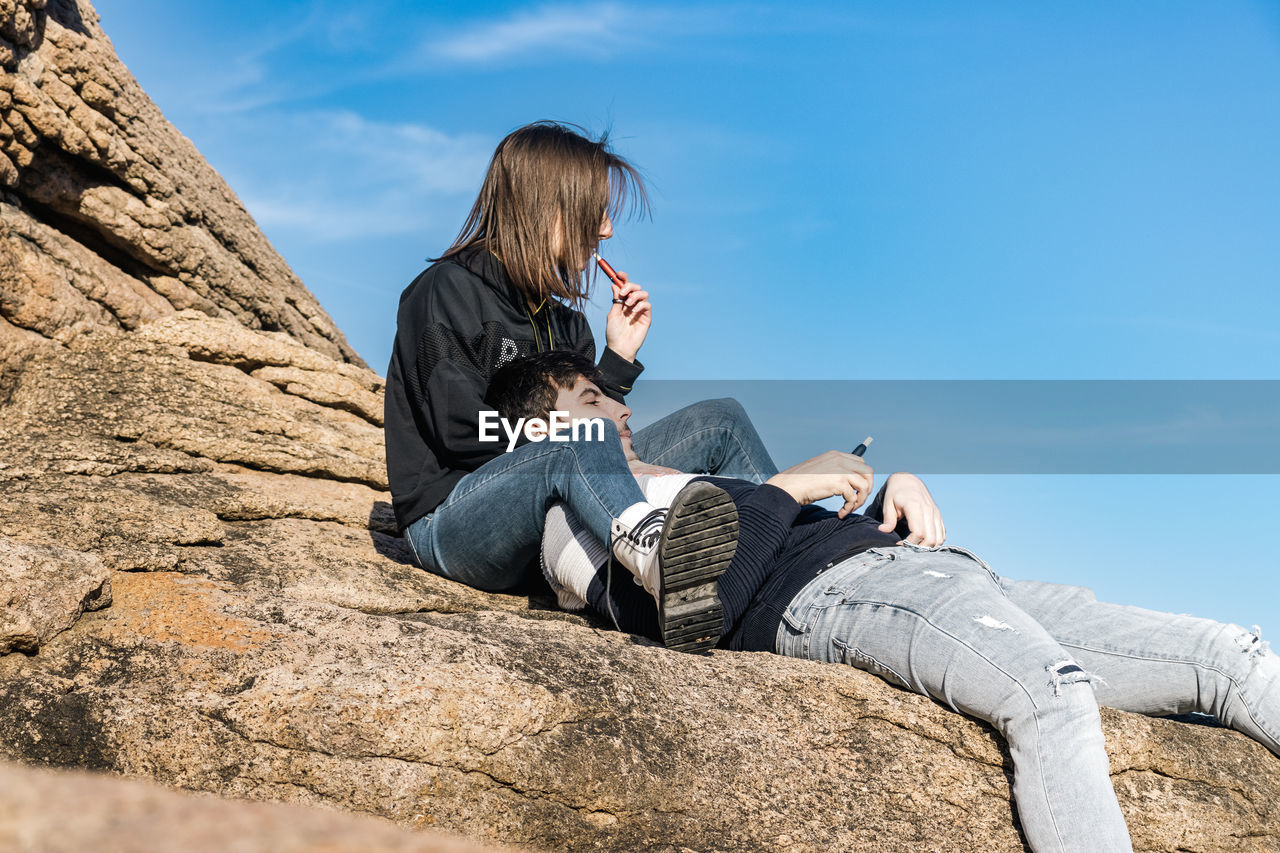 sky, sitting, nature, adult, relaxation, leisure activity, casual clothing, rock, activity, one person, communication, full length, women, technology, wireless technology, lifestyles, day, young adult, reading, outdoors, blue, sunlight, portable information device, photo shoot, mountain, looking, land, holiday, vacation, trip, sunny, person, smartphone, side view, jeans, mobile phone, men, footwear