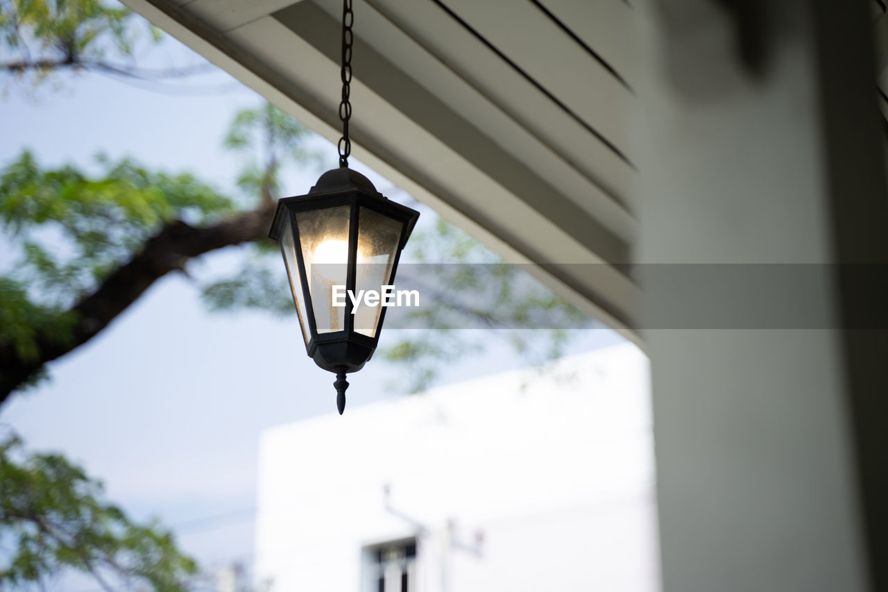 lighting equipment, lighting, hanging, low angle view, electric lamp, architecture, light fixture, lamp, no people, built structure, street light, illuminated, electricity, electric light, lantern, outdoors, street, light, day, light bulb, building exterior, focus on foreground, nature, tree, window, sky, glass