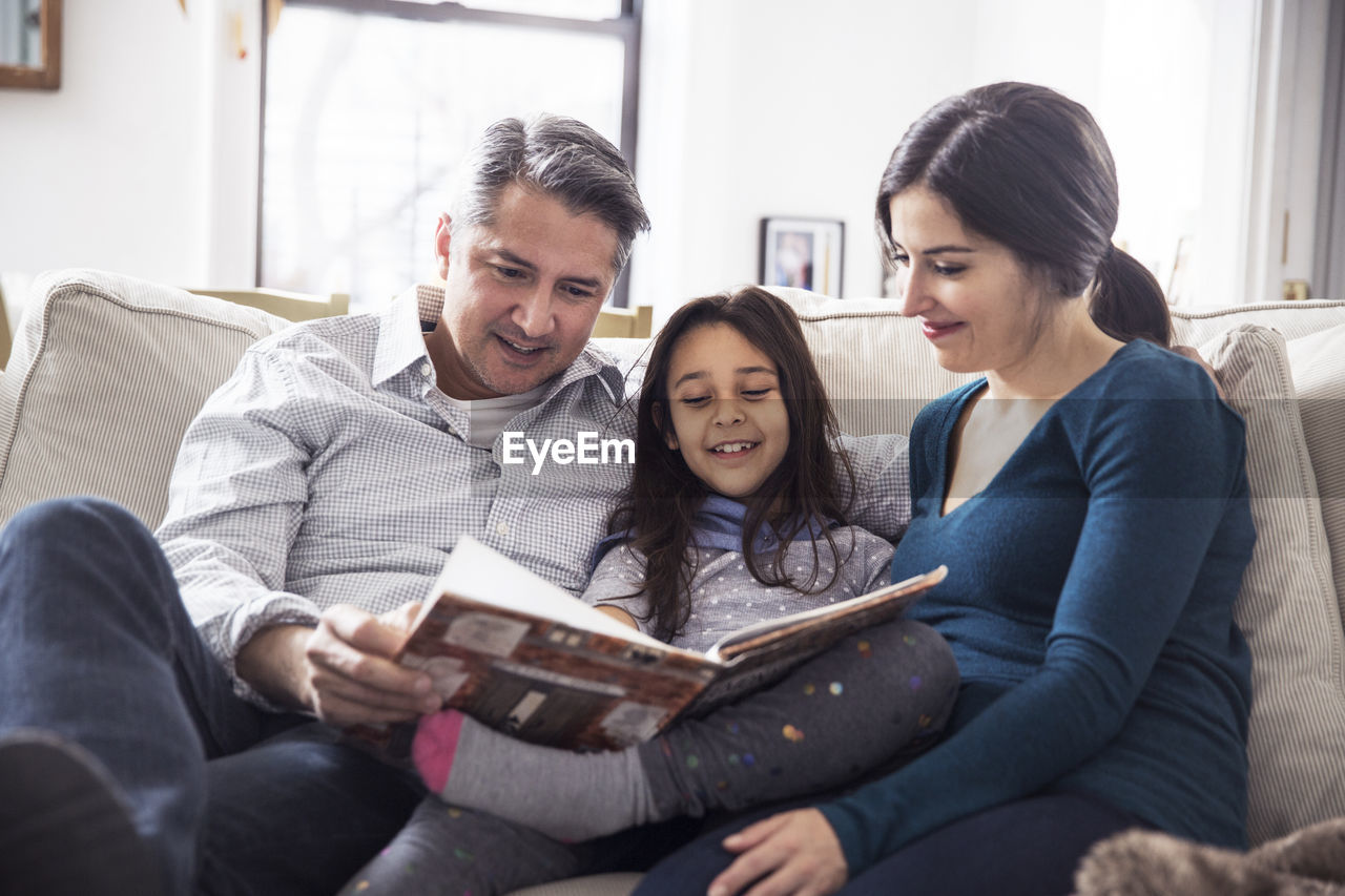 Girl with father and mother reading book while sitting on sofa at home