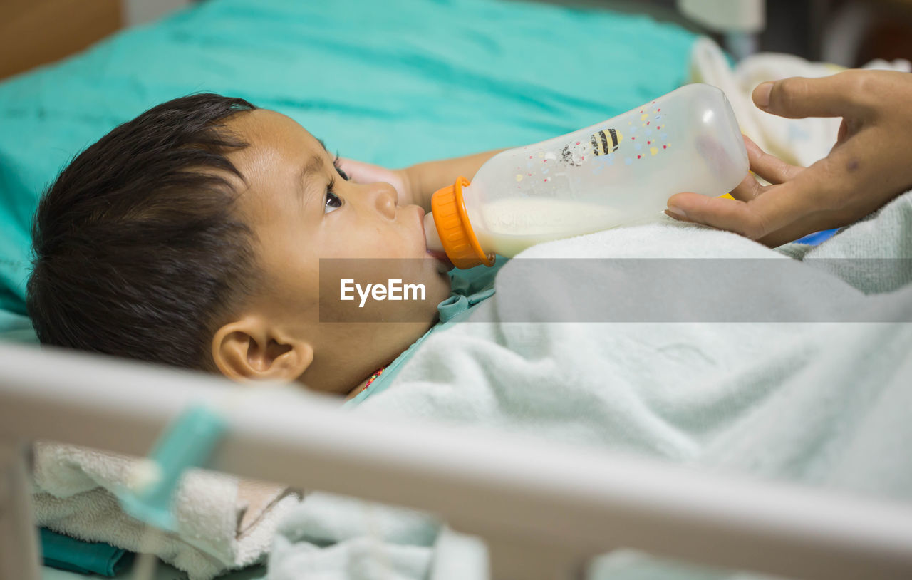 Cropped hands of parent feeding milk to son lying on bed in hospital