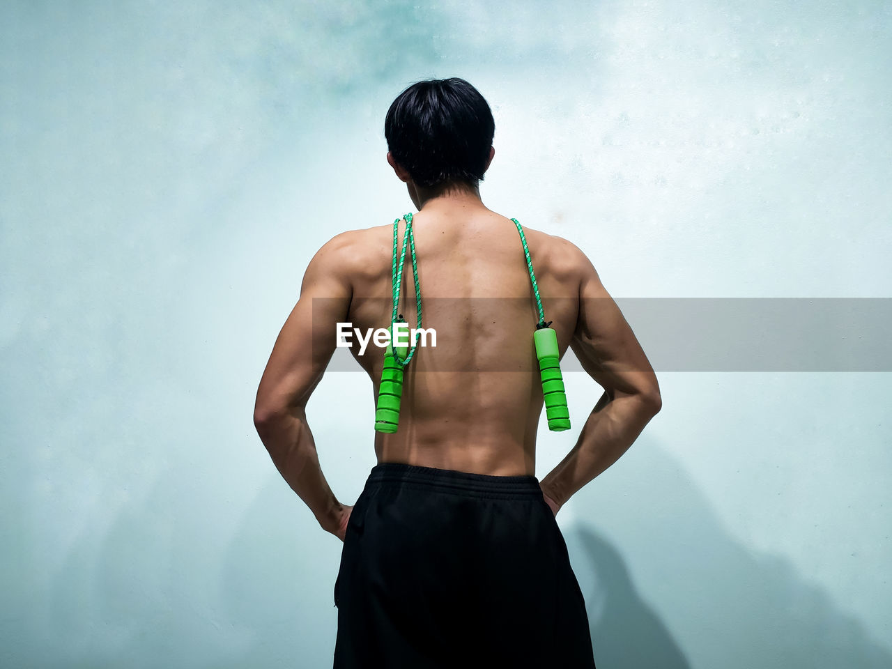 Rear view of shirtless young man with jumping rope standing against wall