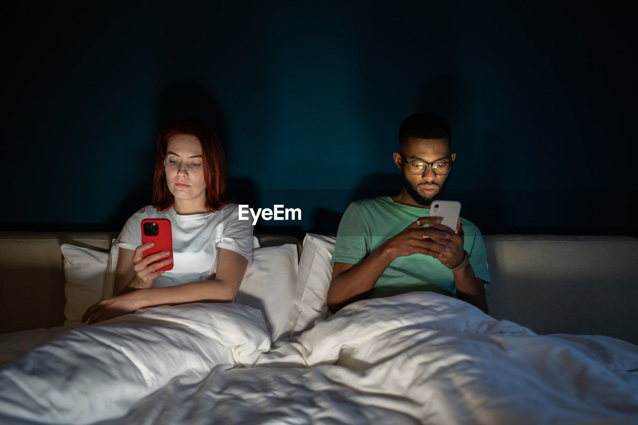Young multiracial couple lying in bed using smartphones not talking
