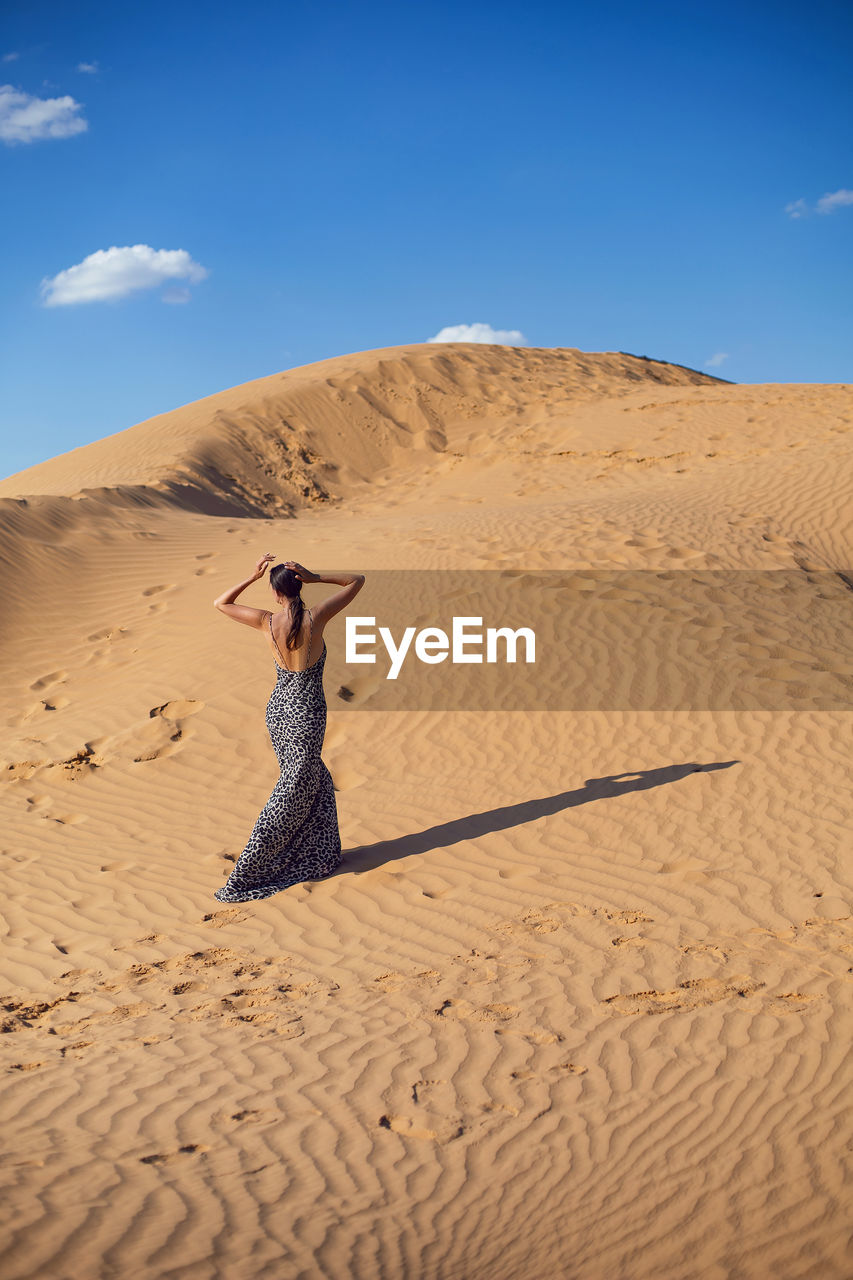 Brunette woman in a long leopard dress stands with her back in the desert at sunset. go everywhere