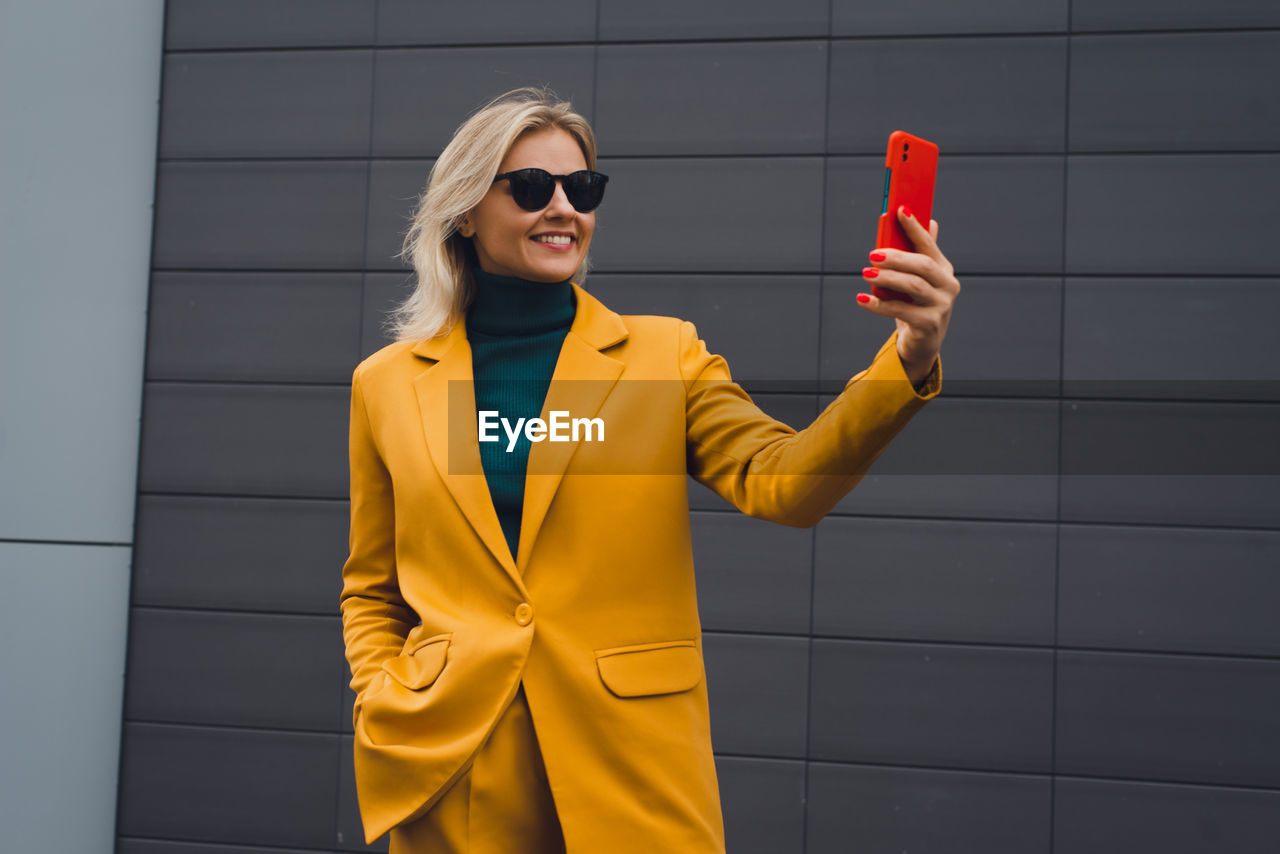 yellow, fashion, adult, glasses, sunglasses, one person, women, smiling, happiness, standing, technology, jacket, young adult, clothing, business, wireless technology, architecture, outerwear, portrait, lifestyles, city, cheerful, spring, communication, coat, blazer, emotion, female, blond hair, person, outdoors, holding, copy space, smartphone, front view, portable information device, sleeve