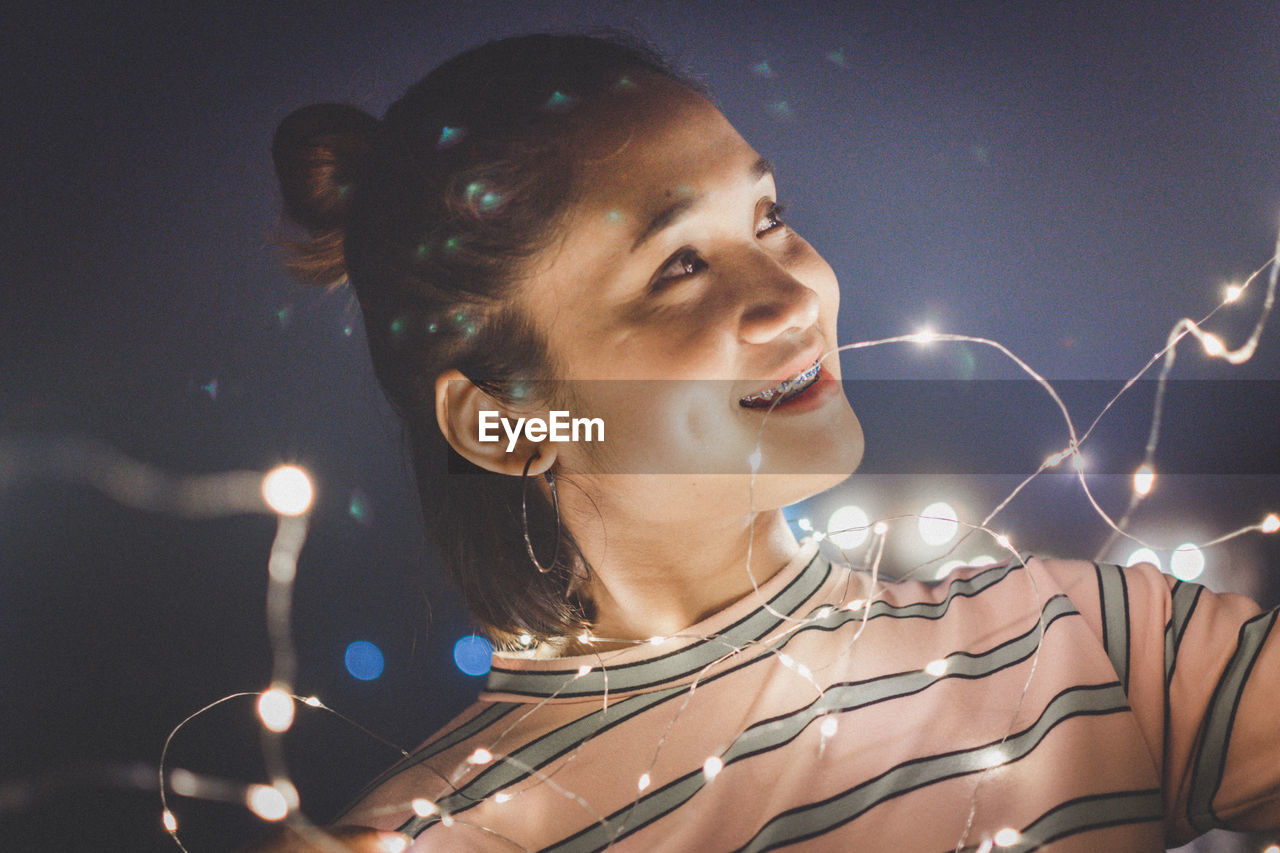 Close-up of smiling young woman with illuminated string lights in city at night