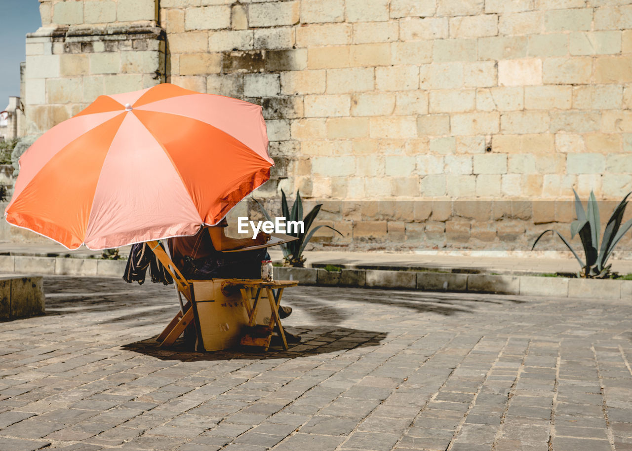 Low section of artist with orange parasol sitting on street against wall