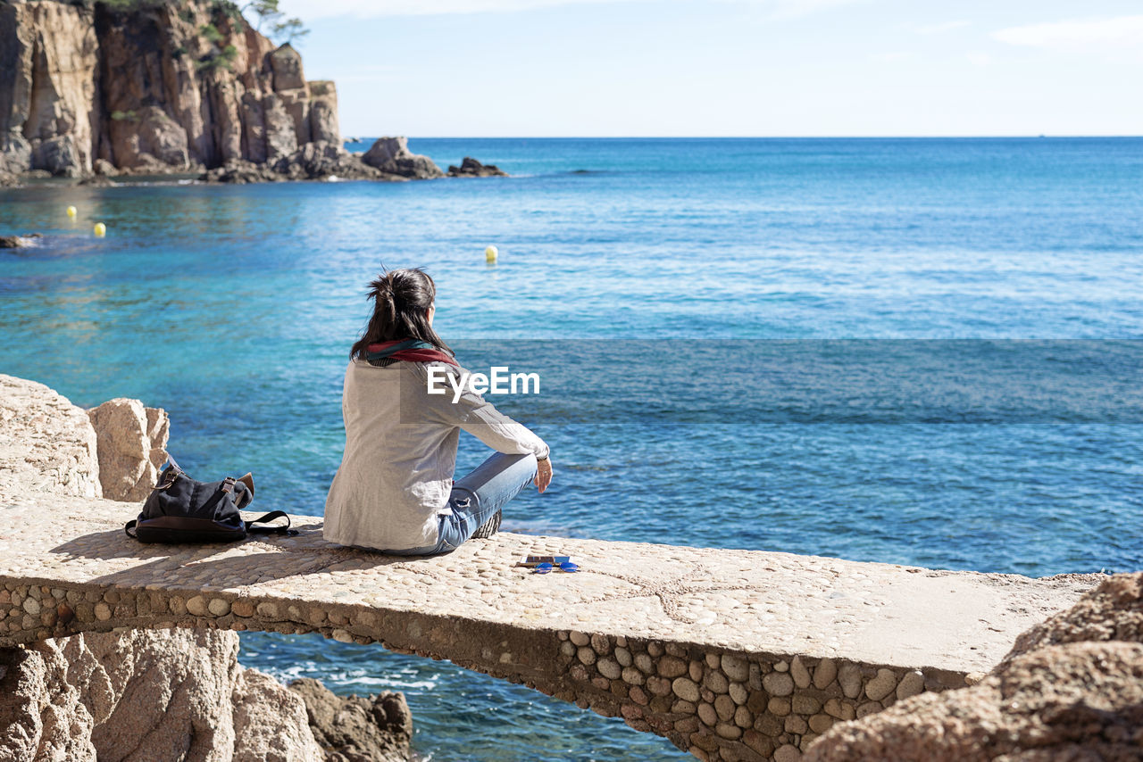 Rear view of a woman relaxing in front of blue sea, sitting on bridge