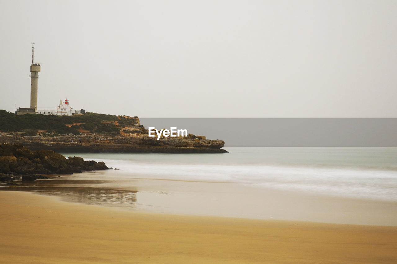 sea, water, lighthouse, beach, land, tower, sky, guidance, architecture, built structure, coast, building exterior, nature, ocean, building, travel destinations, coastline, scenics - nature, horizon, shore, horizon over water, beauty in nature, travel, no people, security, protection, wave, tranquility, breakwater, outdoors, water's edge, clear sky, sand, copy space, tranquil scene, day, environment, tourism, bay, motion, rock