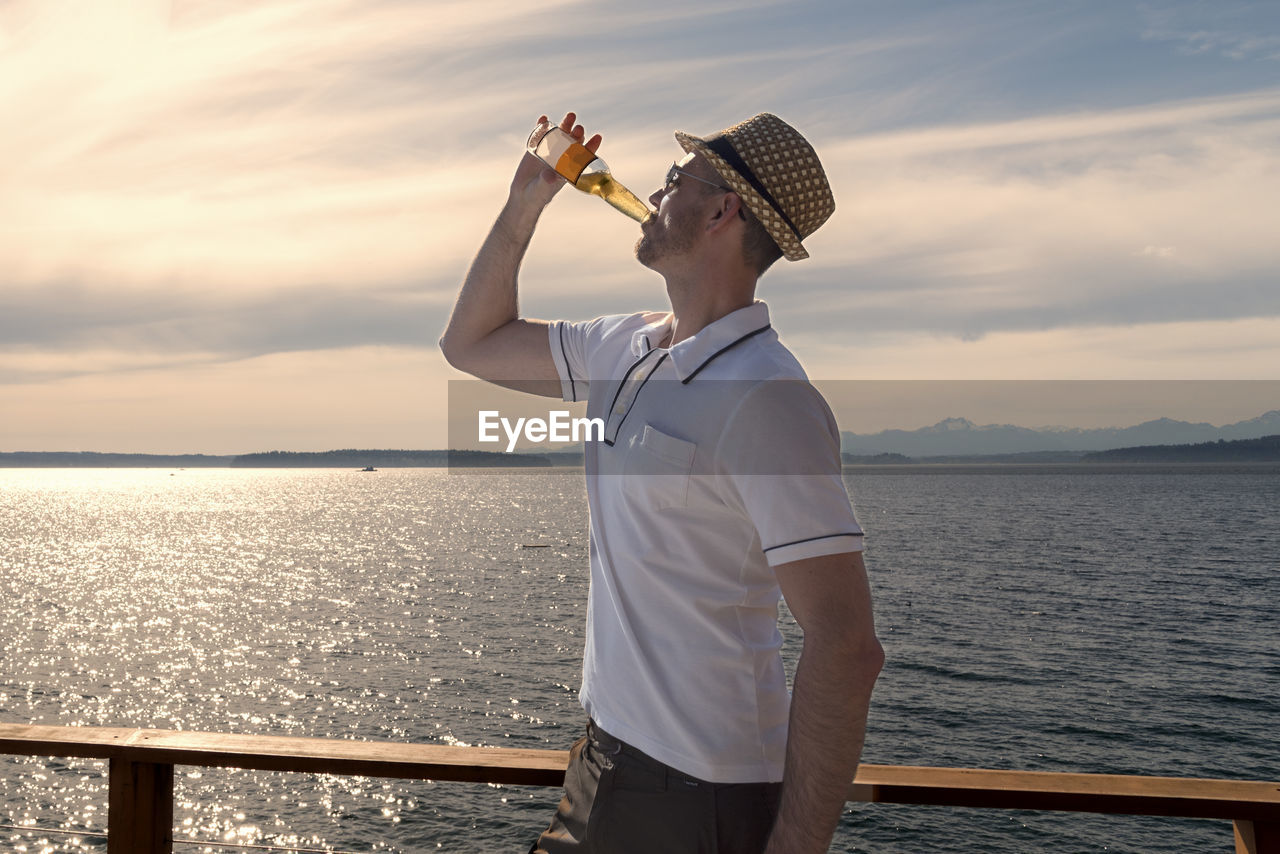 Man drinking beer by sea while wearing hat against sky