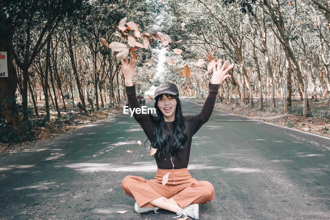 Portrait of smiling young woman throwing leaves while sitting on road amidst trees in forest