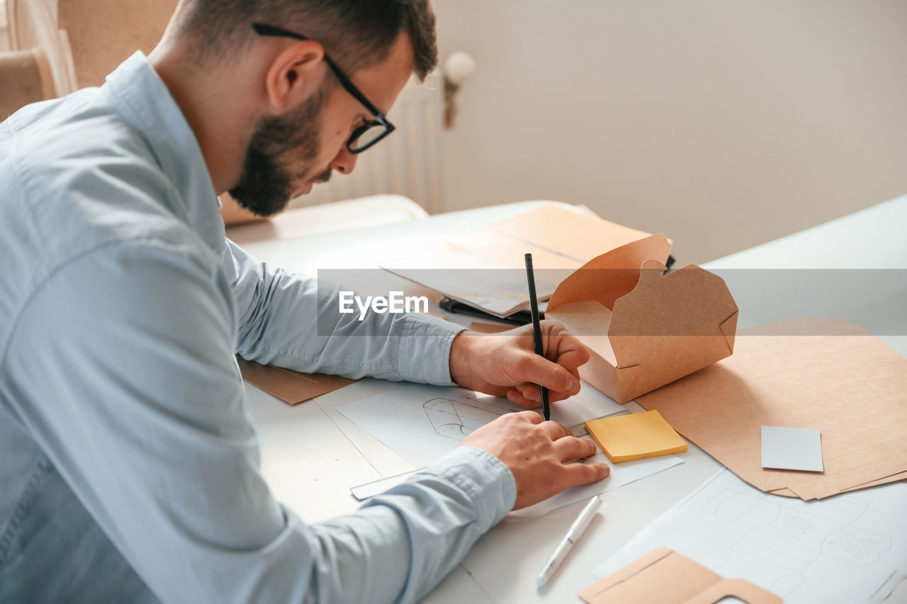 adult, men, indoors, one person, working, business, writing, paper, occupation, business finance and industry, document, person, furniture, table, technology, concentration, office, homework, creativity, looking, beard, learning, new business, home interior, casual clothing, eyeglasses, facial hair, busy, desk, sitting, glasses, finance, businesswear, young adult, looking down, pen, industry, planning, professional occupation