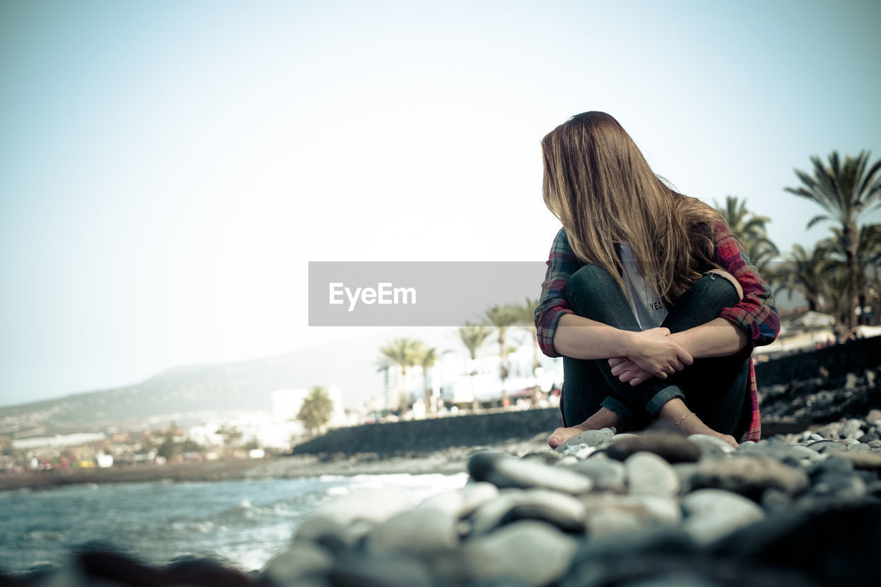 Woman sitting on rock at shore against clear sky