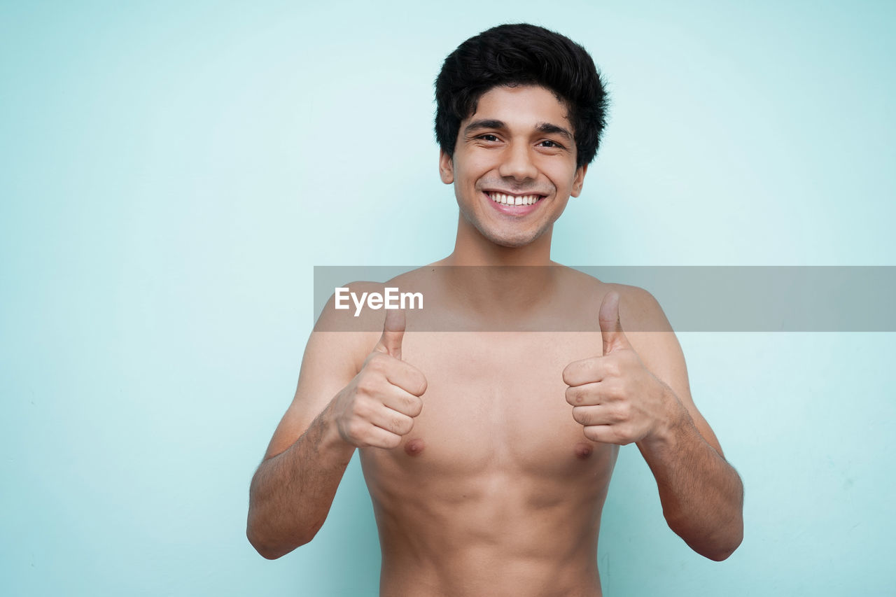 Portrait of smiling shirtless young man showing thumbs up against blue background