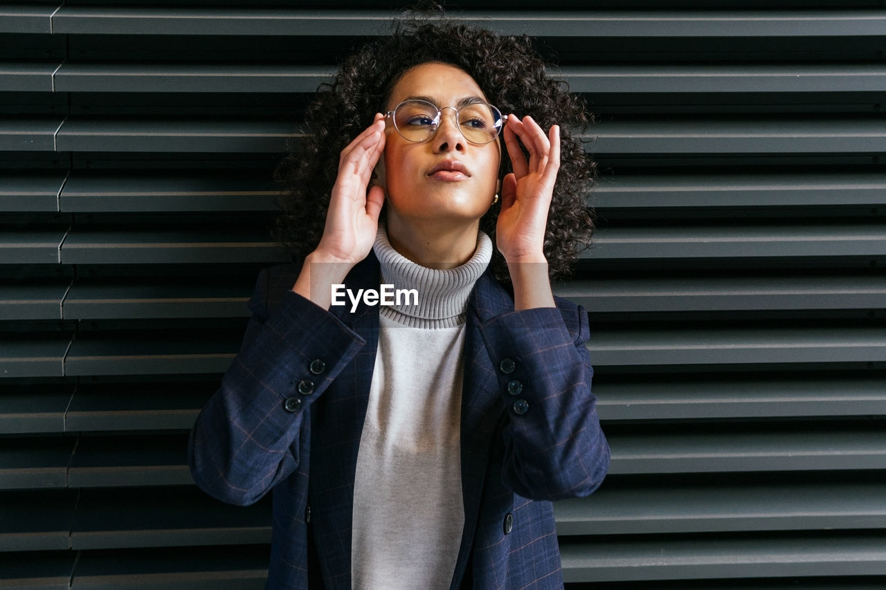 Serious self assured young ethnic female in stylish clothes touching eyeglasses and looking away while standing against gray striped wall