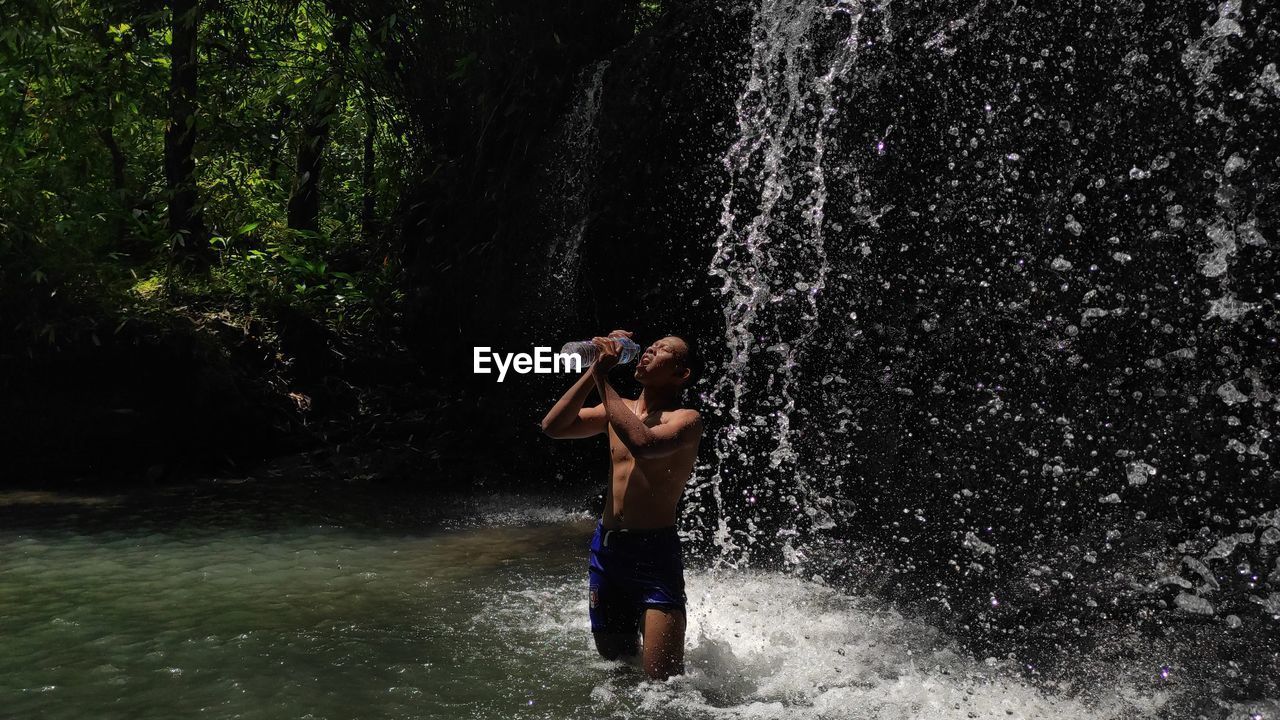 Shirtless young man drinking water from bottle while standing by waterfall