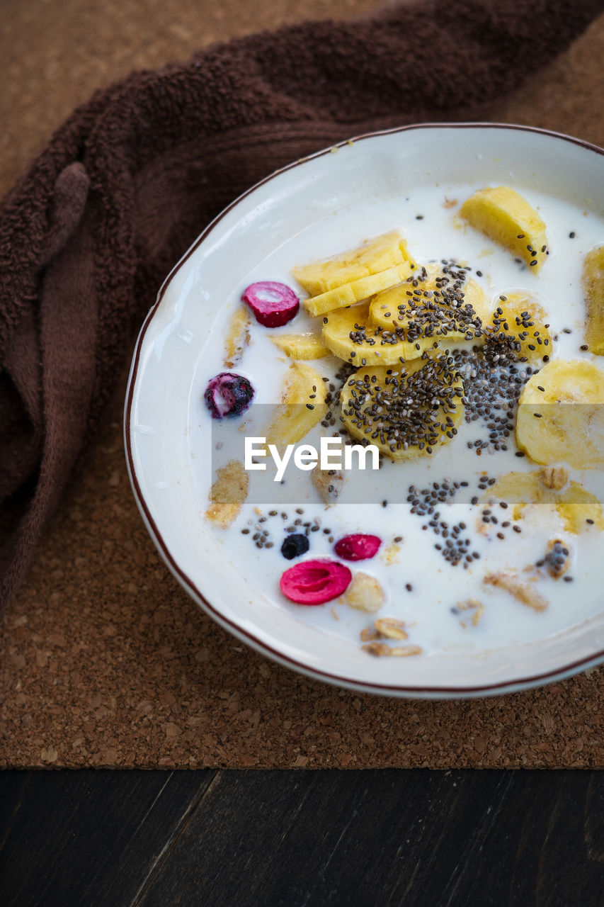 Fresh granola, banana with milk in a white bowl on brown background. sprinkled with chia seeds.