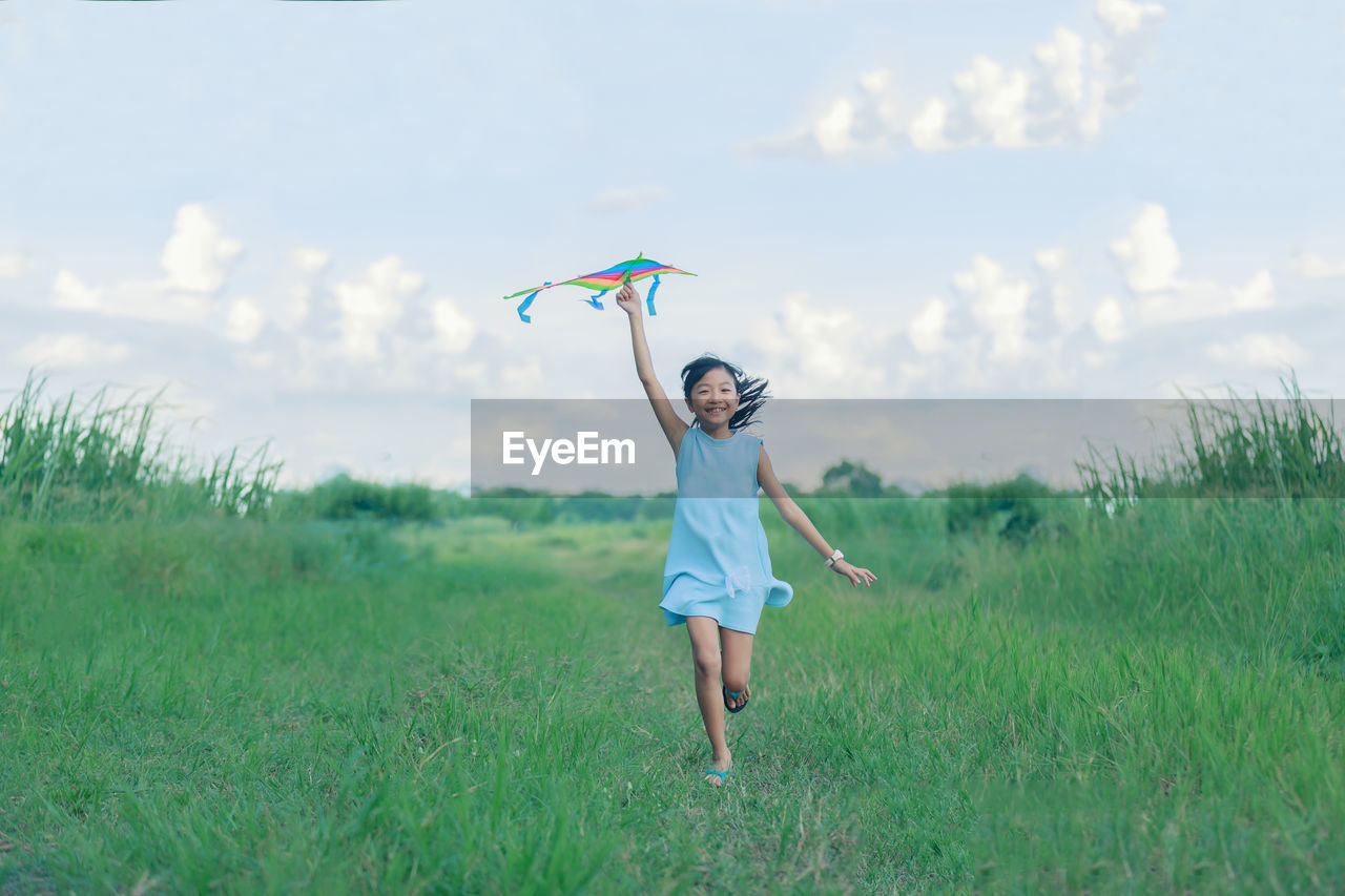 Full length of happy girl holding kite while running on grassy field against sky