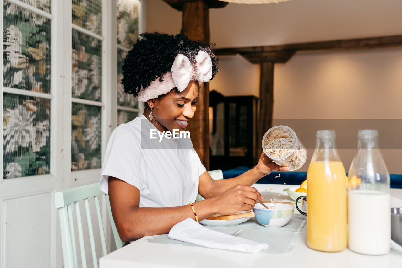 Happy african american lady smiling and adding fresh oatmeal in bowl while sitting at table and having breakfast at home