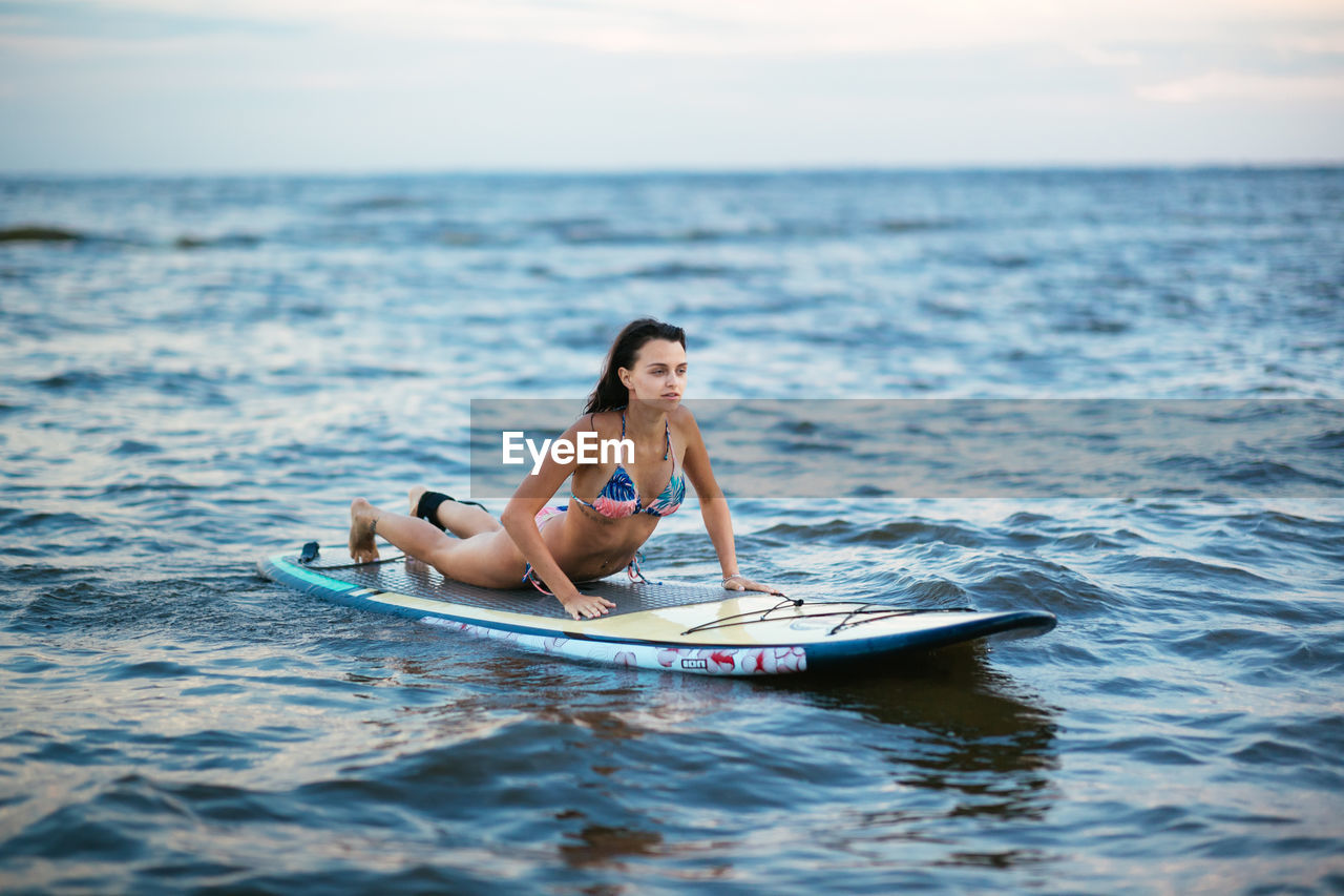 Woman in bikini lying over surfboard on sea against sky