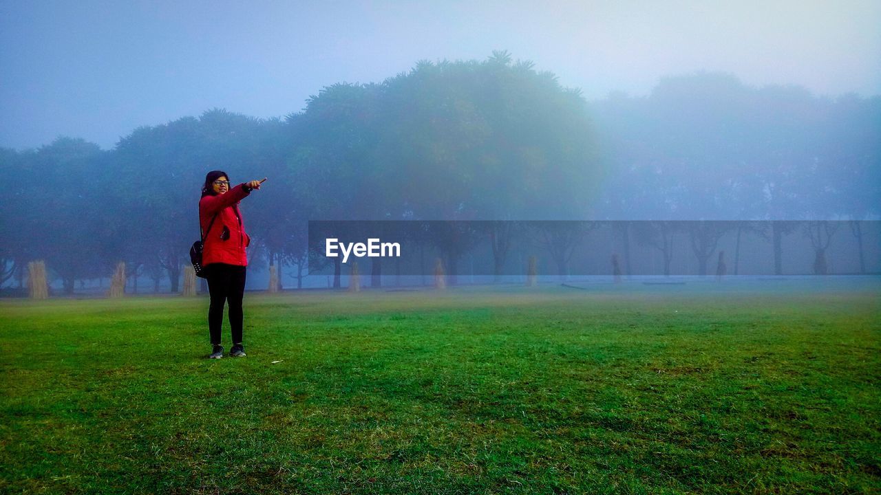 Full length of young woman pointing while standing on grassy field during foggy weather