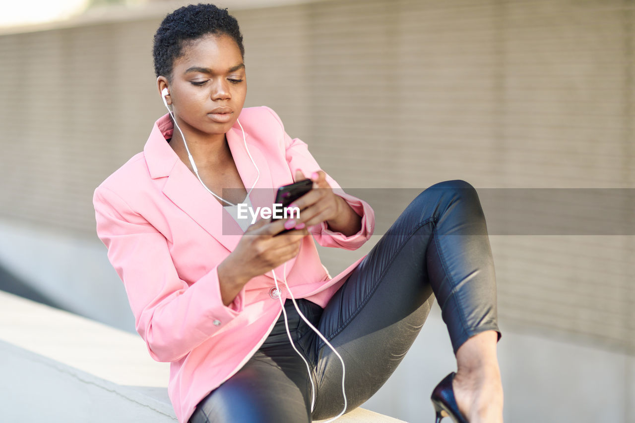 Full length of young woman listening music while sitting on retaining wall outdoors