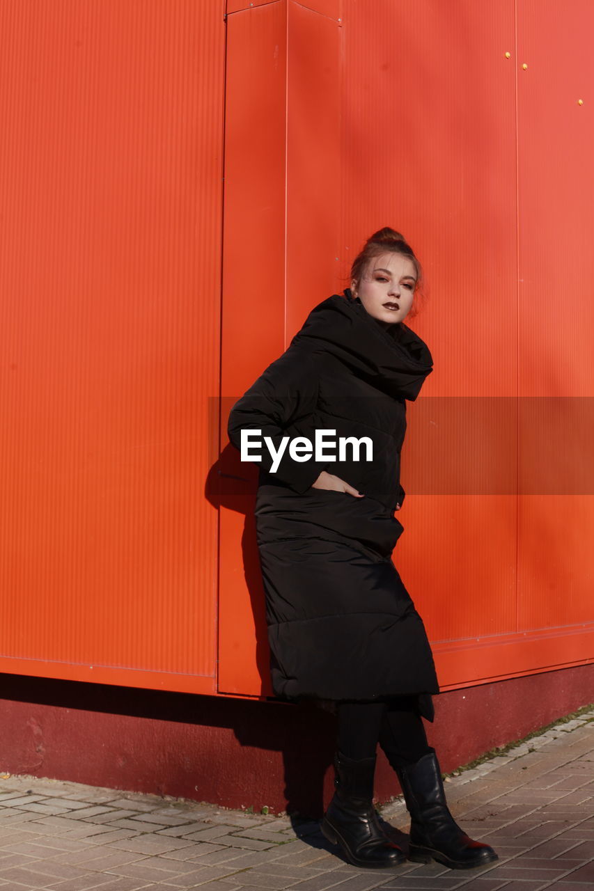 Fashionable teenage girl wearing black warm clothing standing against red wall during sunny day