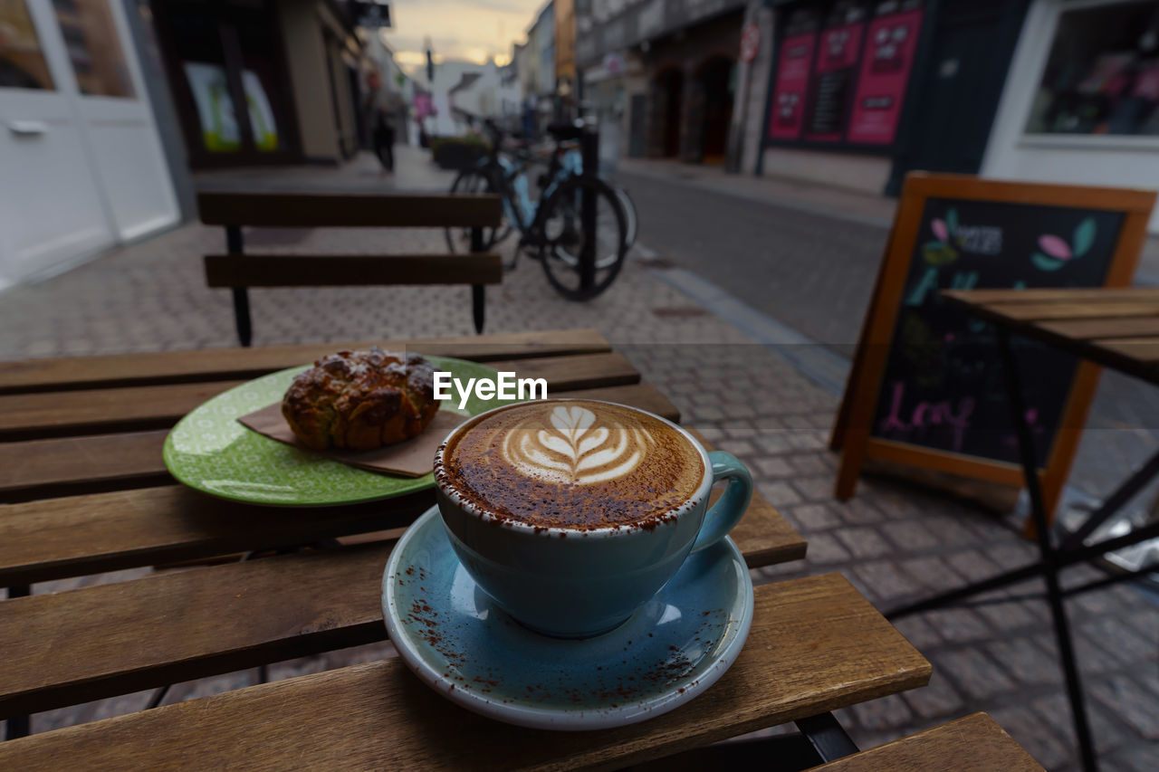 food and drink, bicycle, coffee, drink, table, cup, mug, food, coffee cup, refreshment, freshness, sweet food, hot drink, coffeehouse, cafe, vehicle, dessert, sweet, wheel, cappuccino, land vehicle, baked, frothy drink, furniture, no people, bicycle wheel, still life, saucer, crockery, focus on foreground, indoors, wood, architecture, chocolate, kitchen utensil, day, latte