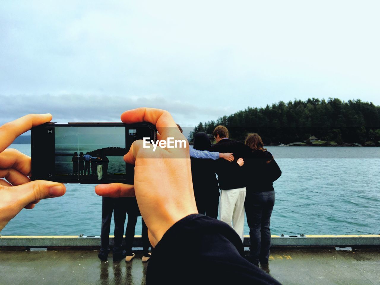 Cropped image of hands photographing friends by lake against sky