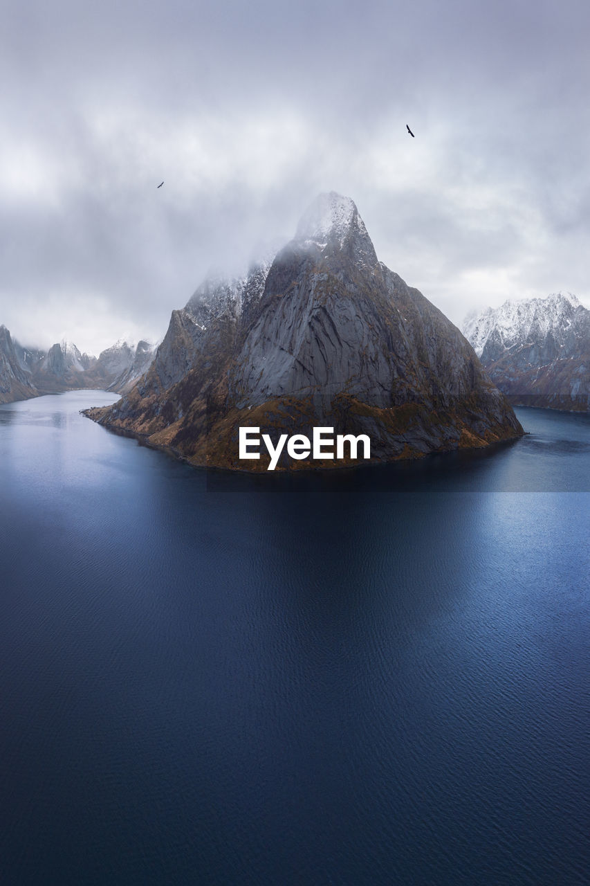 Aerial view of calm water of sea washing island with rocky formations covered in snow under cloudy sky