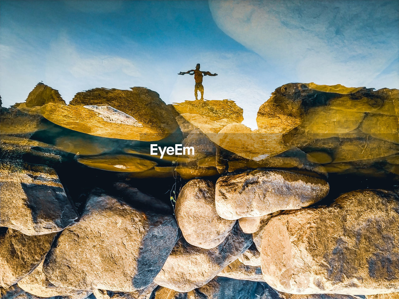 MAN STANDING ON ROCK BY WATER AGAINST SKY