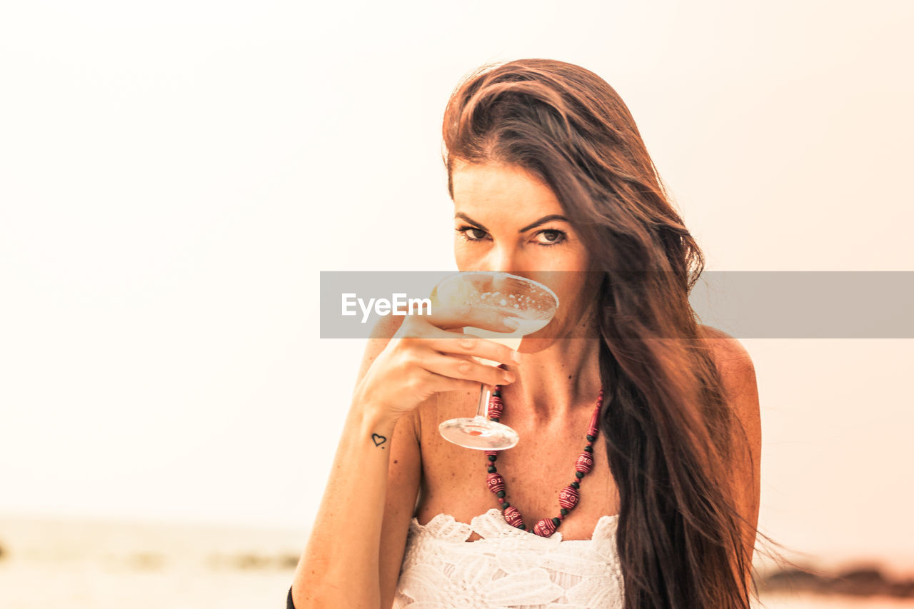 Portrait of mature woman holding drink at beach against sky