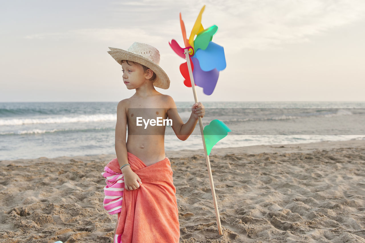 Boy wearing hat holding windmill toy while standing at beach