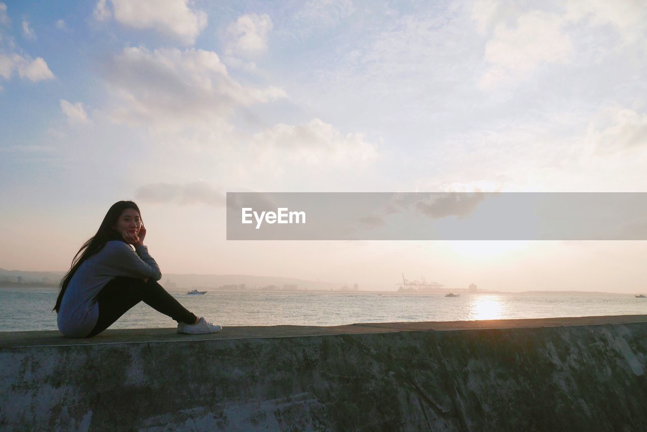 Young woman sitting on retaining wall against sea at beach