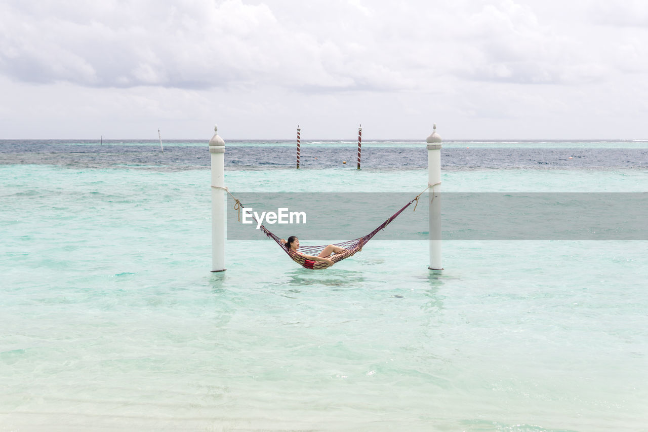Woman in red swimsuit lying in hammock swing over ocean surf line relaxing in maldives on cloudy day