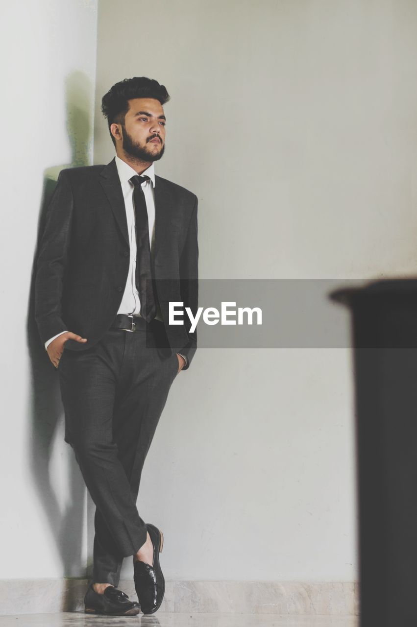 Full length of handsome young man with hands in pockets standing against wall at home