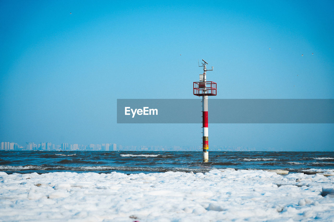 sky, ocean, tower, sea, blue, guidance, water, nature, snow, architecture, beach, wave, cold temperature, clear sky, built structure, land, no people, winter, lighthouse, horizon, scenics - nature, day, beauty in nature, communication, outdoors, security, copy space, tranquility, wind, protection, environment, building exterior, building, travel destinations, cloud, frozen, tranquil scene, horizon over water, sign, travel