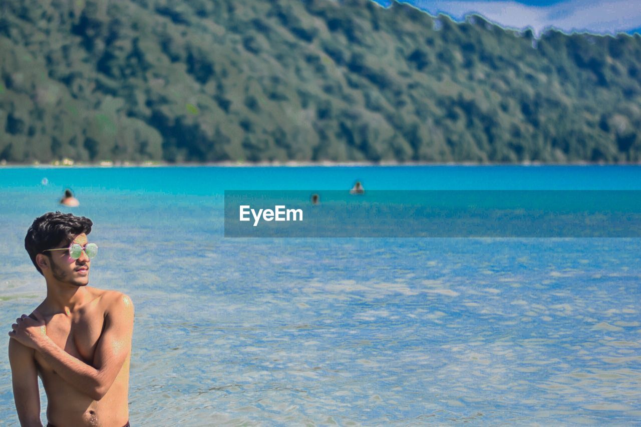 Shirtless man wearing sunglasses while standing at beach against mountain