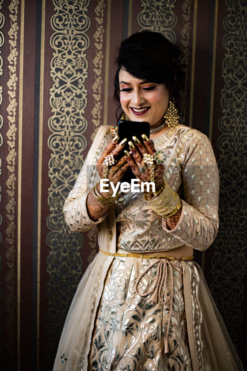 Smiling woman using phone while standing against curtain