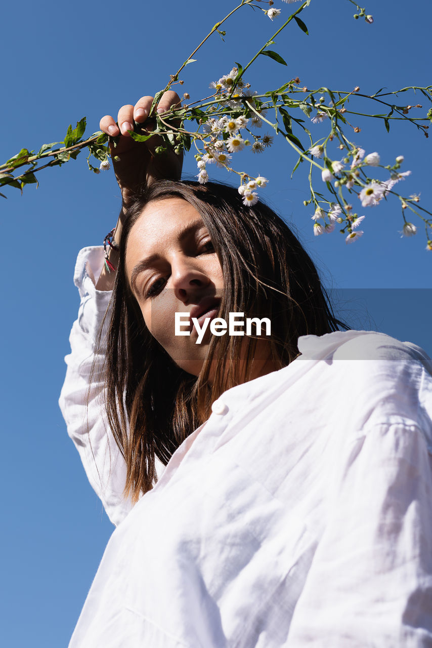 Low angle portrait of young woman holding flowers against clear sky