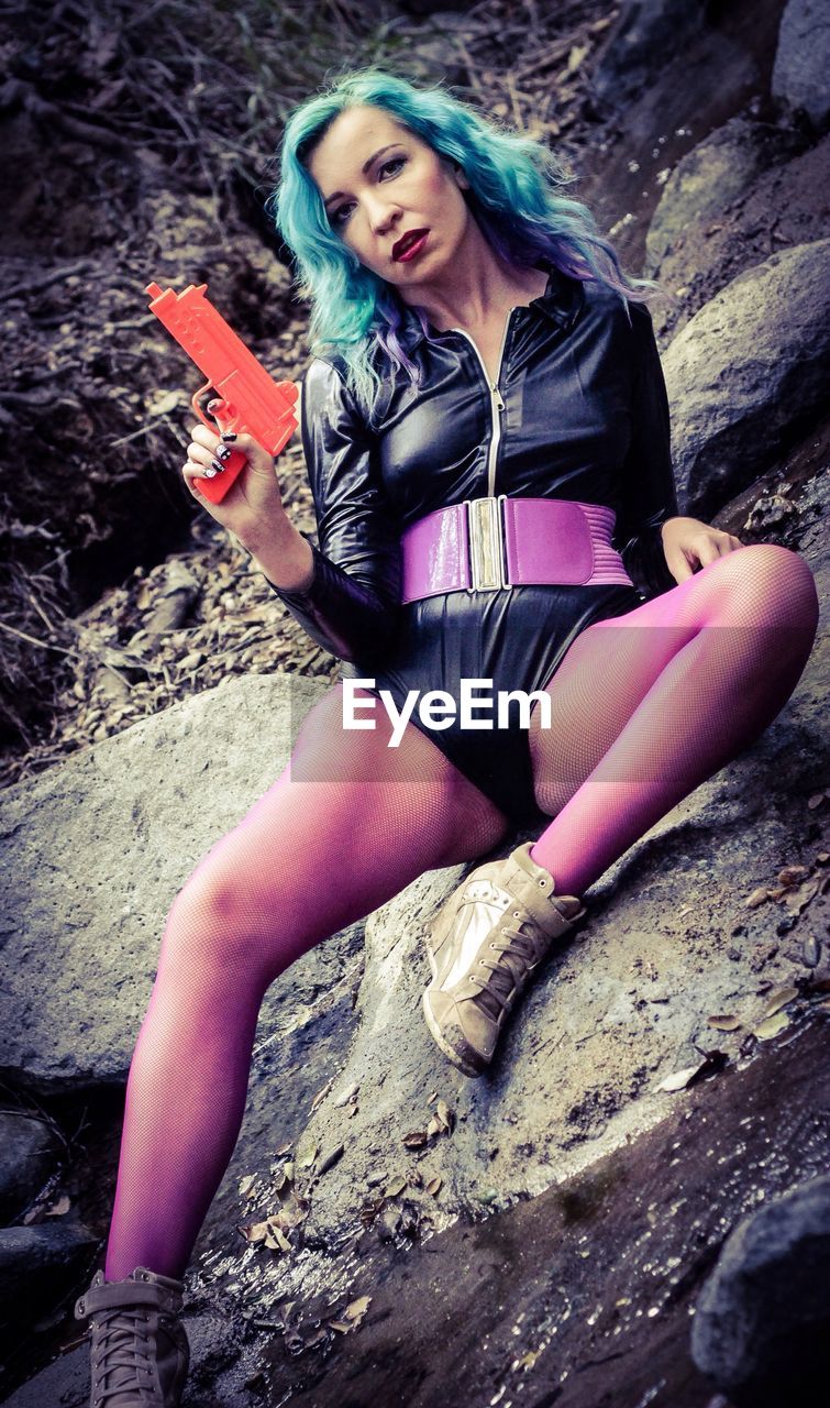 Full length portrait of female model holding toy gun while sitting on rock