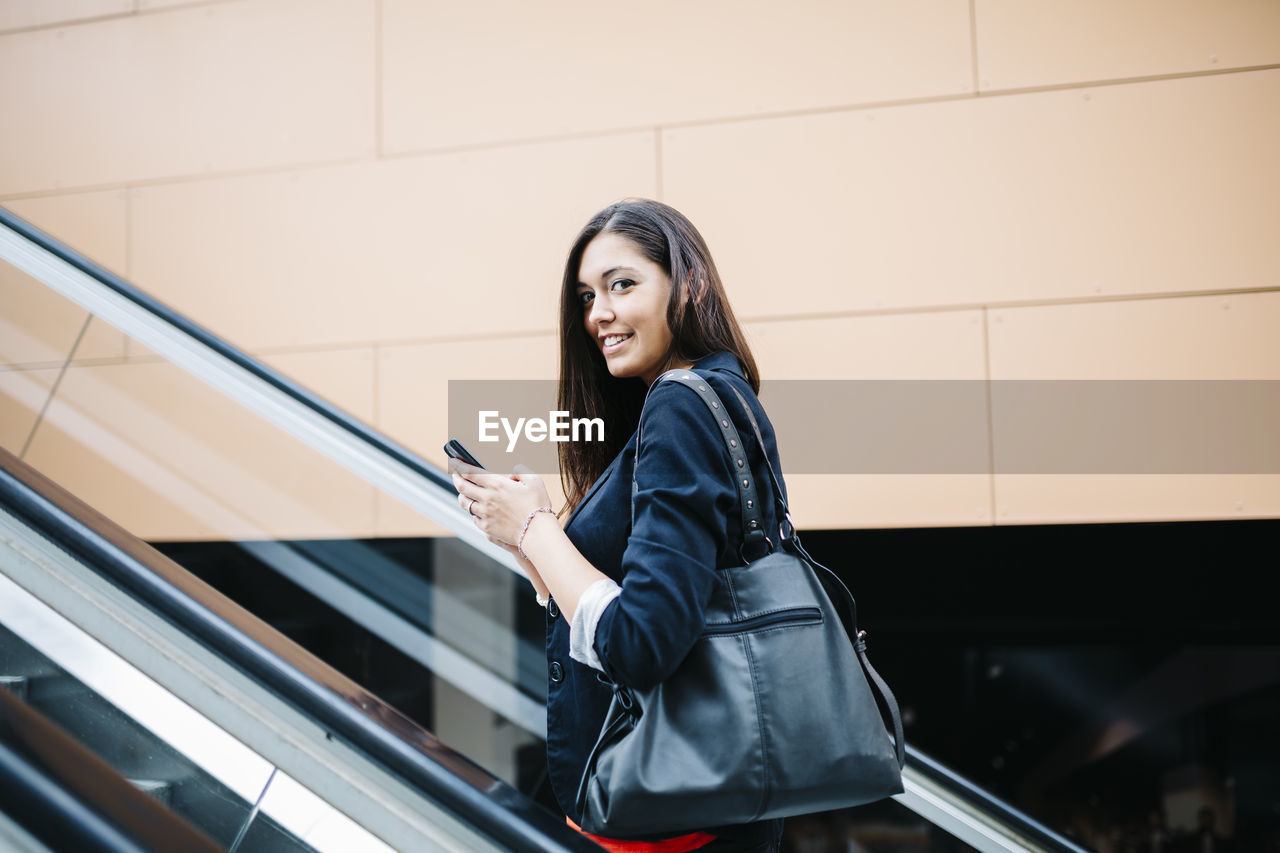 Portrait of smiling young woman using mobile phone while standing on escalator