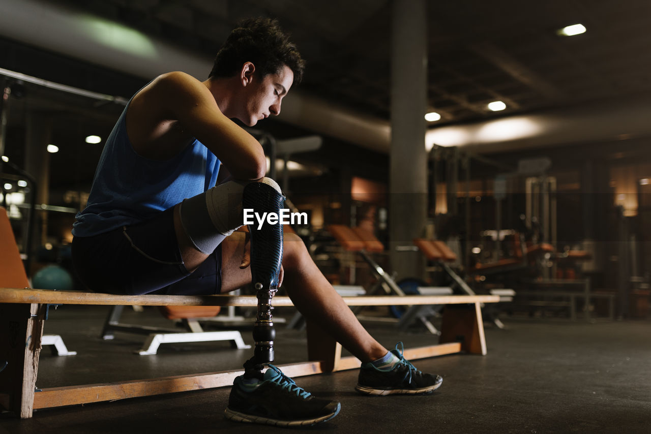 Full length of young man with prosthetic leg sitting on bench in gym