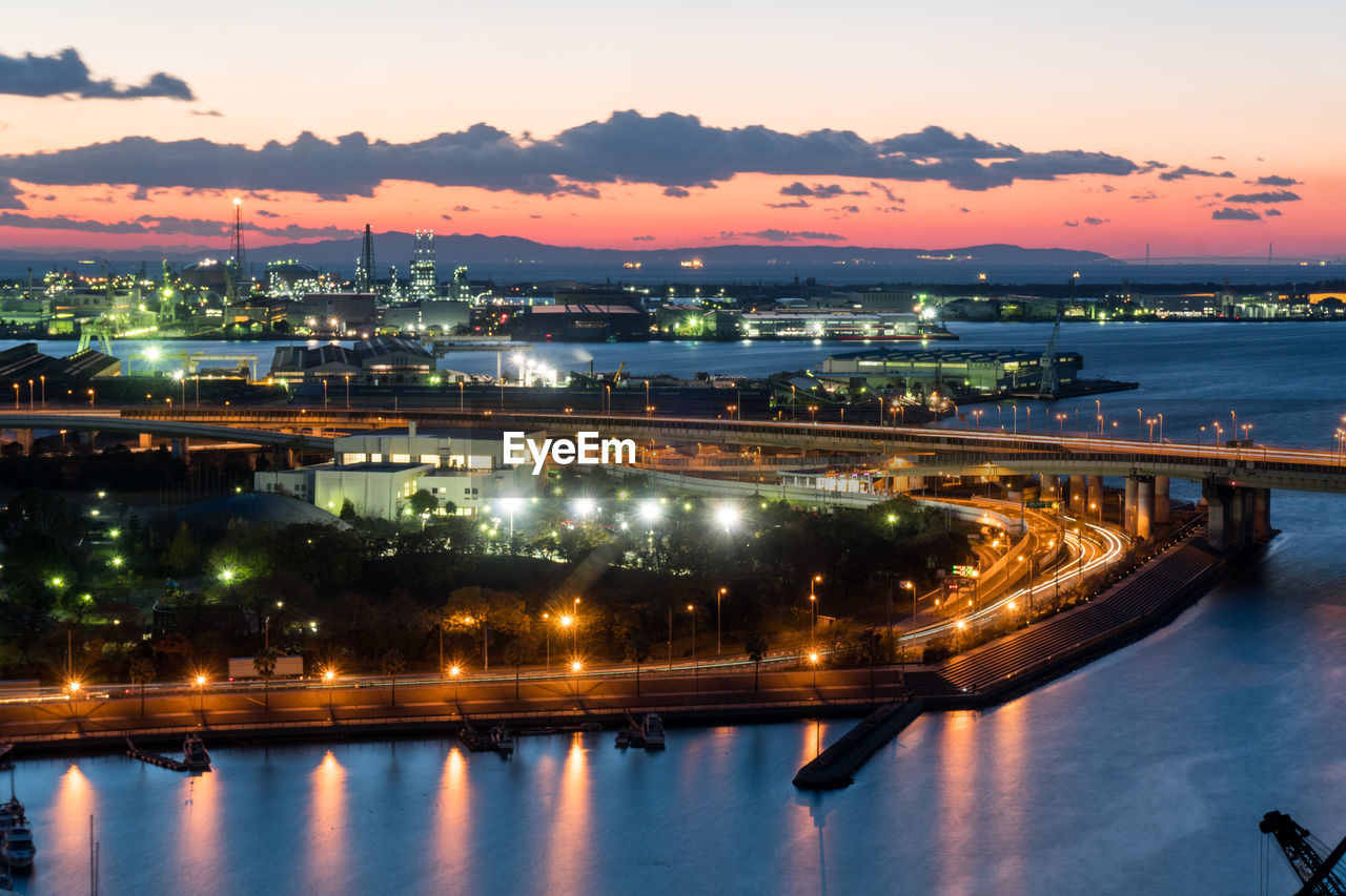 Illuminated city by river against sky at sunset