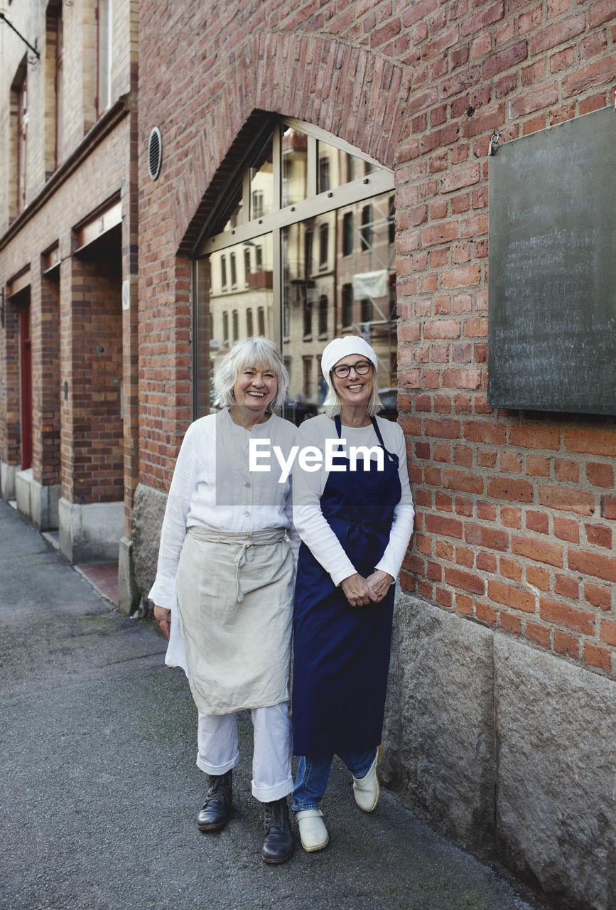 Portrait of smiling female bakers standing by brick wall outside bakery