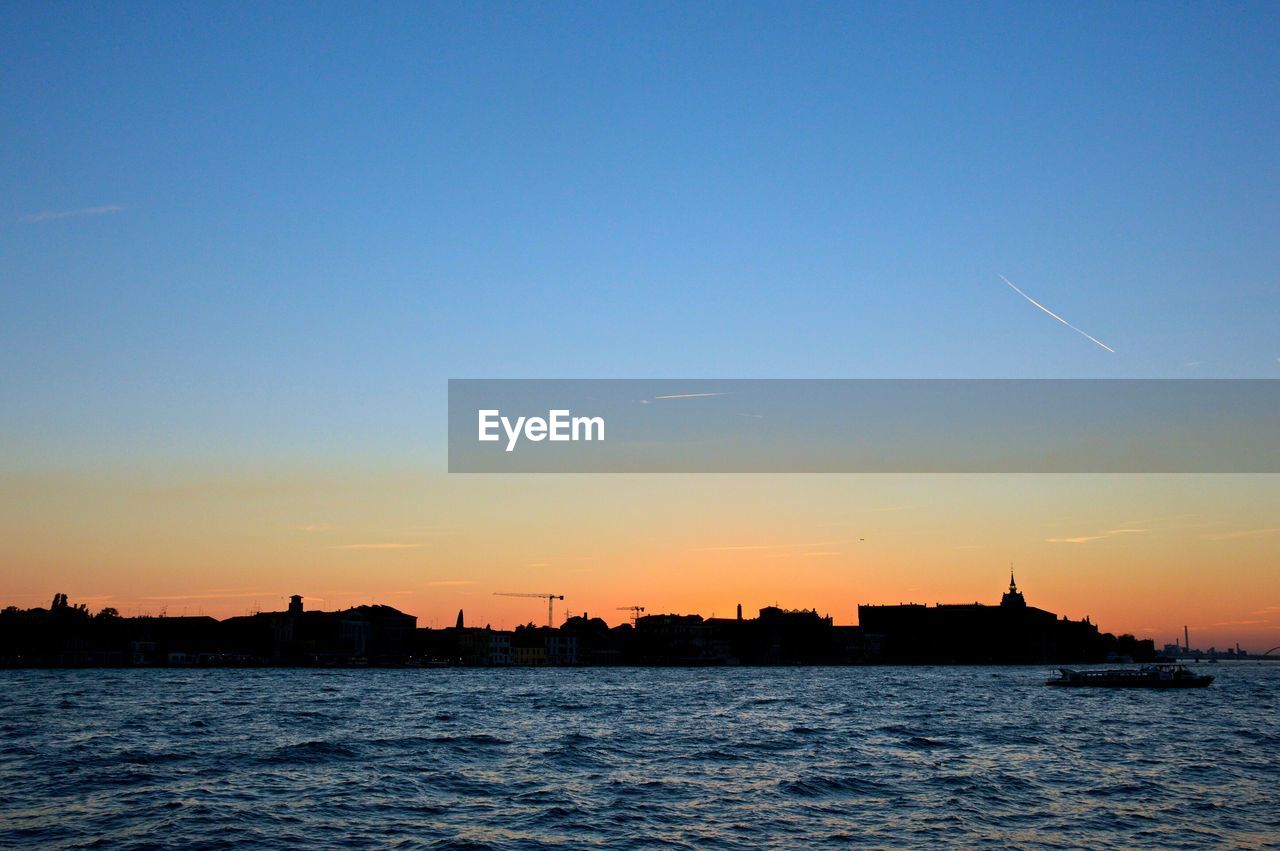 Sea by silhouette city against clear sky during sunset